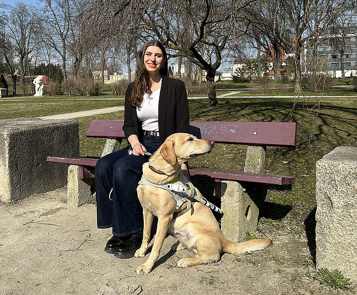 Eine junge Frau mit sehr langen braunen Haaren blickt lächelnd in die Kamera, zu ihren Füßen vor der Parkbank ein Führhund.