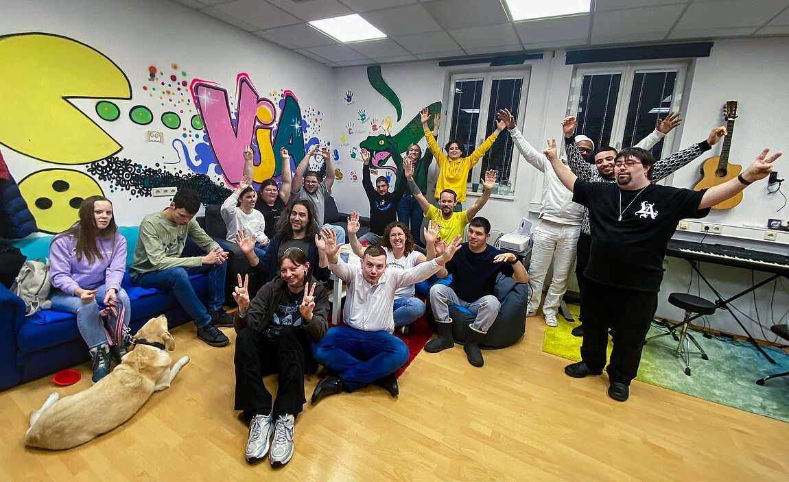 17 Personen stehen oder sitzen, teilweise am Boden, im Jugendraum. Viele heben für das Foto die Arme nach oben, die gute Stimmung im Raum ist zu spüren. Im Vordergrund links liegt auch ein Hund am Boden.