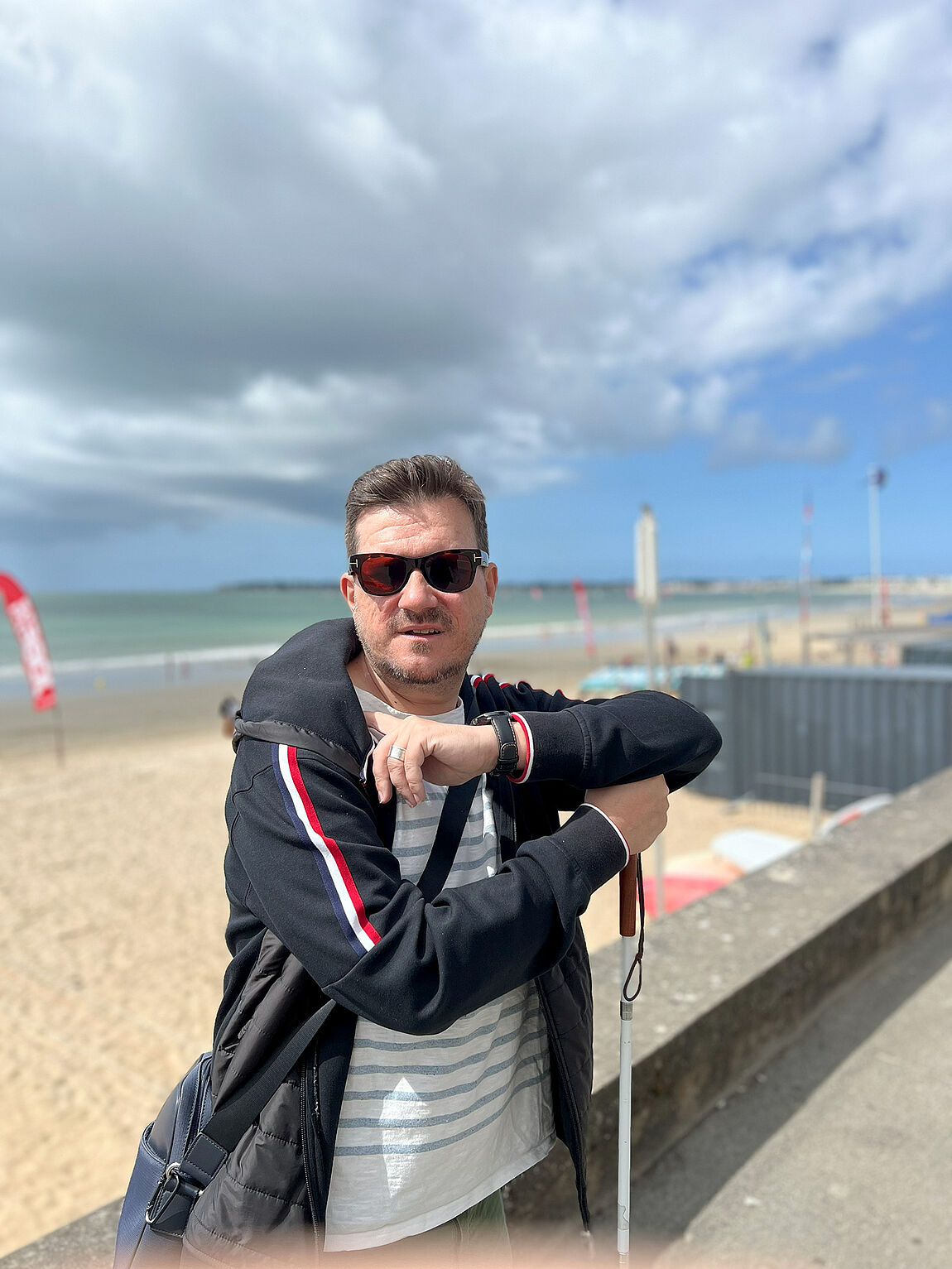 An einer Strandpromenade lehnt ein Mann mit Windjacke an einer niedrigen Mauer, den Arm lässig auf einen Weißen Stock gestützt.