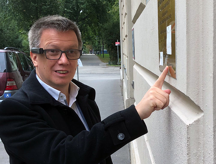 Ein Mann mit Brille inklusive Kameraaufsatz steht vor einem Arztschild an einer Außenwand, er zeigt mit dem Finger auf das Schild.
