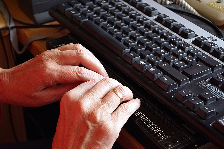 Die Finger zweier Hände gleiten über die elektronische Braillezeile, dahinter ist eine Computer-Tastatur zu sehen.