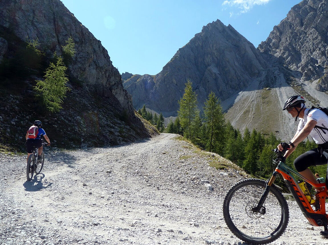 Zwei Mountainbiker auf einer Schotterstraße zwischen hohen Berghügeln.