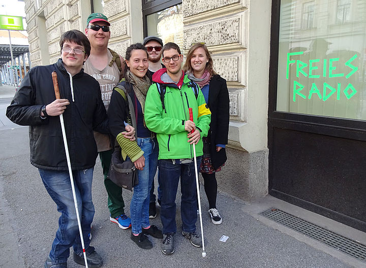 Sechs Jugendliche, darunter zwei mit weißem Stock, stehen als Gruppe lachend vor einer Fensterfront mit der Aufschrift "Freies Radio"