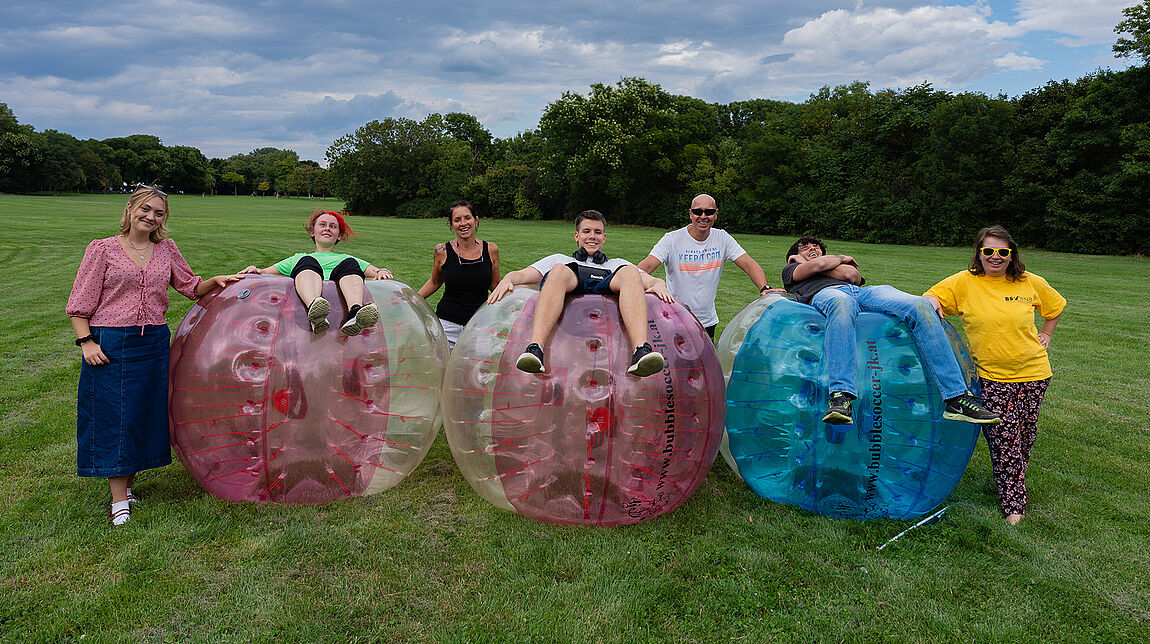 Auf drei aufgeblasenen Riesenbällen auf einer Wiese liegen ebensoviele Jugendliche. Vier Personen des Veranstaltungs- bzw. Jugendteams stehen bei ihnen für ein Gruppenfoto.