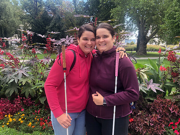 Zwei junge Frauen umarmen sich seitlich und lachen dabei in die Kamera, jede von ihnen hält einen weißen Stock. Hinter ihnen viele Blumen in einem Park.