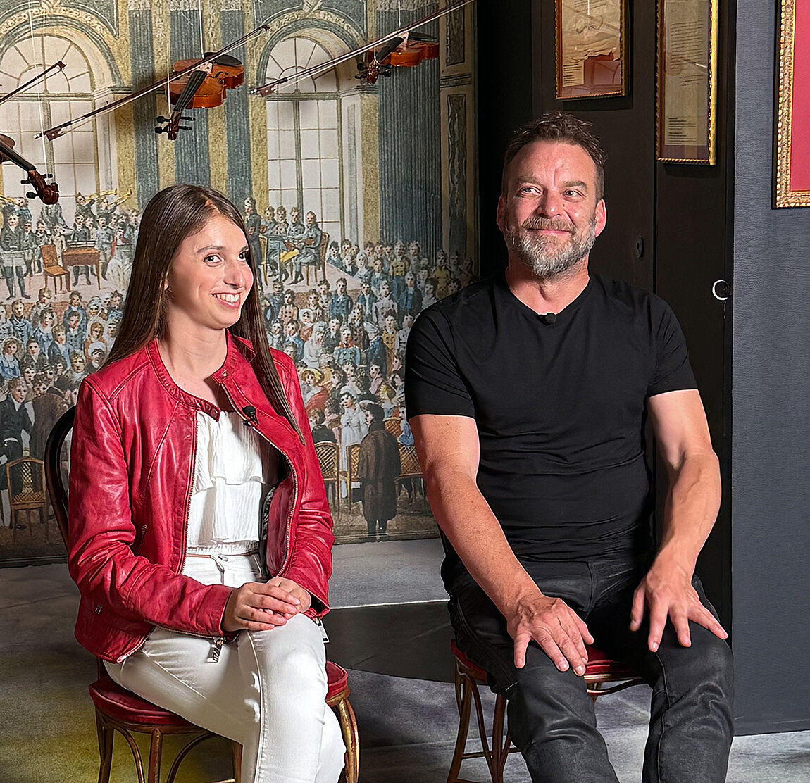 Die Sängerin und ihr Produzent sitzen entspannt auf zwei Stühlen vor einem Wandbild, das ein klassisches Konzert darstellt.