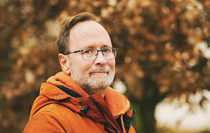 Ein Mann mittleren Alters mit Brille und kurzem Bart. Er steht in einem herbstlichen Park.
