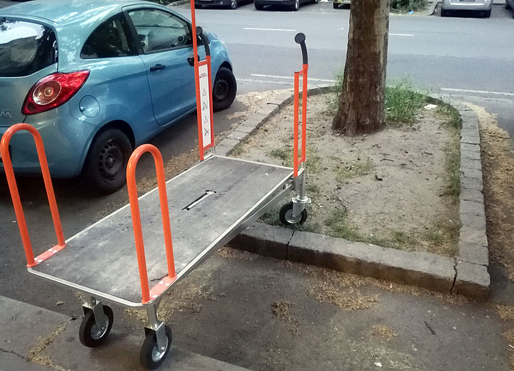 Ein massiver Einkaufswagen eines Baumarktes mit großer Aufladefläche und hochragenden Griffstangen an allen vier Ecken ragt vom Gehsteig hinüber auf eine Bauminsel zwischen parkenden Autos.