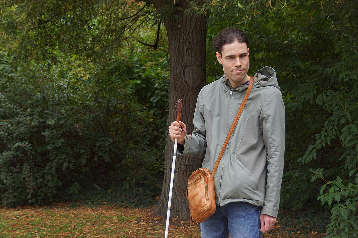 Ein junger Mann mit Umhängetasche und weißem Stock vor einem Baum in einem Park.