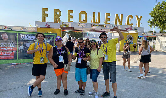 Zusammen mit dem VJA-Team strahlen zwei junge Erwachsene in die Kamera und zeigen Daumen hoch, direkt vor dem riesigen Frequency-Logo im Eingangsbereich des Musikfestivalgeländes.