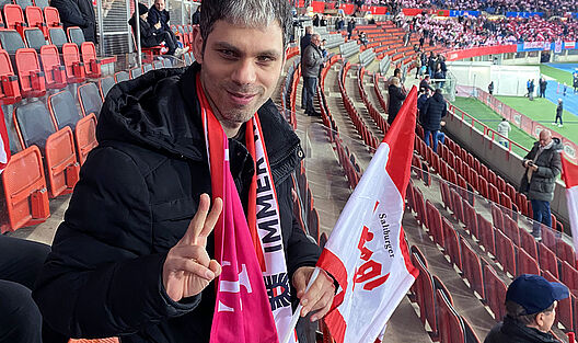 Ein junger Mann befindet sich auf den Tribünen des Ernst-Happel-Stadions hält eine österreichische Flagge in der Hand und trägt einen rot-weiß-roten Fanschal während er in die Kamera lacht. Hinter ihm sind der Spielrasen sowie mehrere Sitzreihen des Stadions sichtbar.