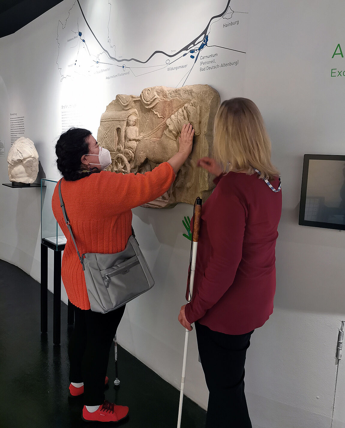 Zwei Frauen stehen vor einer Wand mit Relief, eine der Frauen ertastet mit den Händen Details.