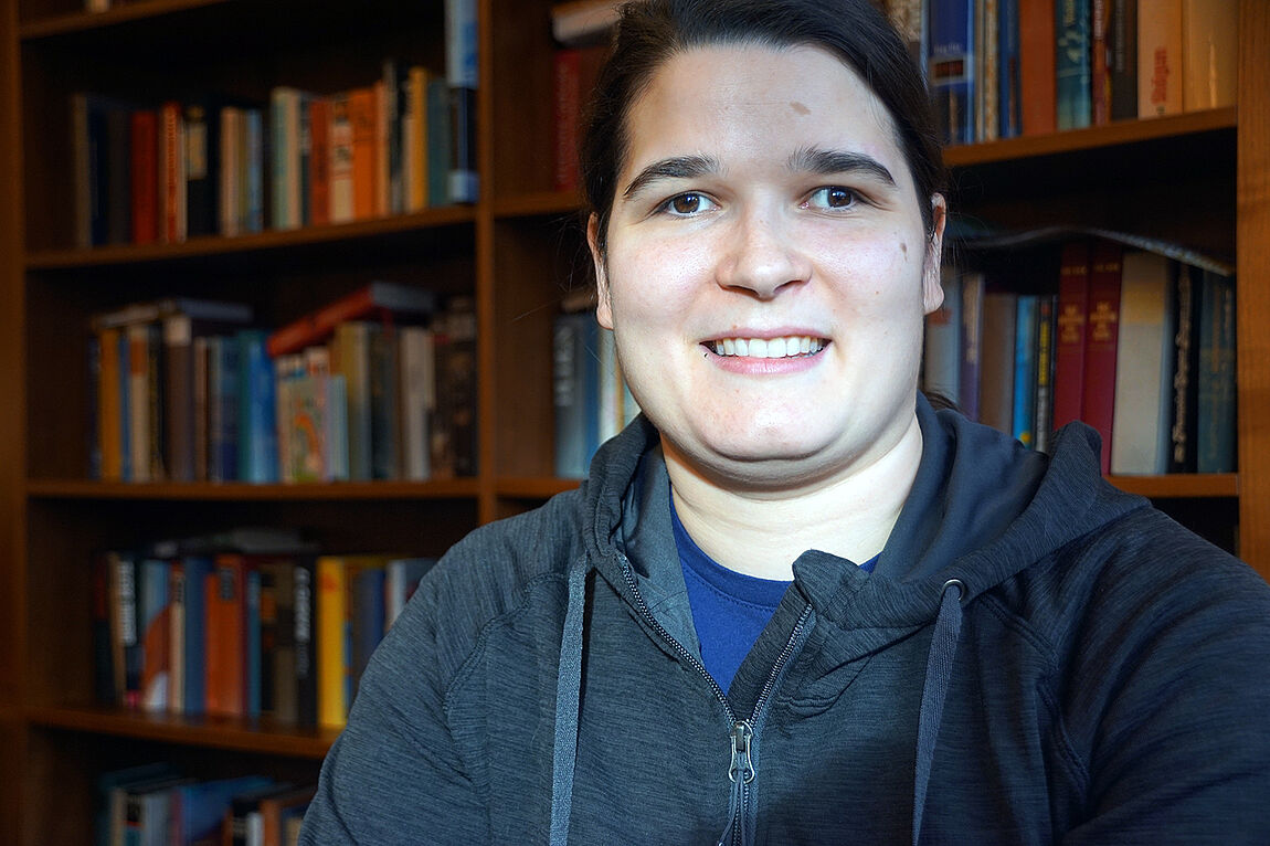Portraitaufnahme einer jungen Frau, im Hintergrund Bücherregale.