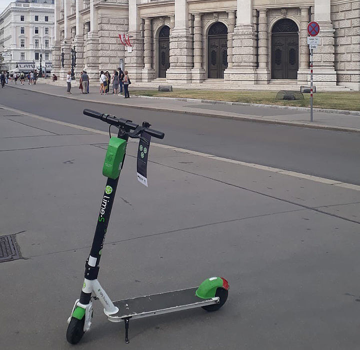 Ein E-Scooter steht auf einem breiten Gehsteig, dahinter ist das Burgtheater zu erkennen. An der Lenkstange des Scooters hängt ein Informations-Flyer von LIME