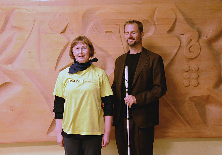 Vor einem großen Wandrelief im Foyer des Louis Braille Hauses: Ein Mann mit Weißem Stock und geschlossenen Augen berührt den Ellenbogen einer Frau die neben ihm steht und sich bereit macht, ihn so zu führen.