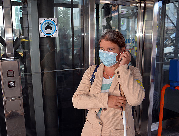 Eine Frau vor einem Aufzug hinunter zum Bahnsteig hält mit der einen Hand den Weißen Stock, während sie mit der anderen Hand eine MNS-Maske über ihr Ohr fädelt. Am Aufzugschacht ein Aufkleber mit einem schemenhaft dargestellten dunklen Kopf mit heller Maske und der Aufschrift: Bitte Mund und Nase bedecken!