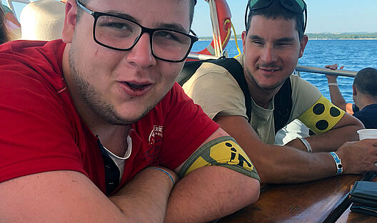Zwei junge Männer lächeln in die Kamera. Sie sitzen auf Holztischen an Bord eines großen Schiffs, während im Hintergrund das weiße Segel sowie das Meer vor der Küste Kroatiens zu sehen ist.