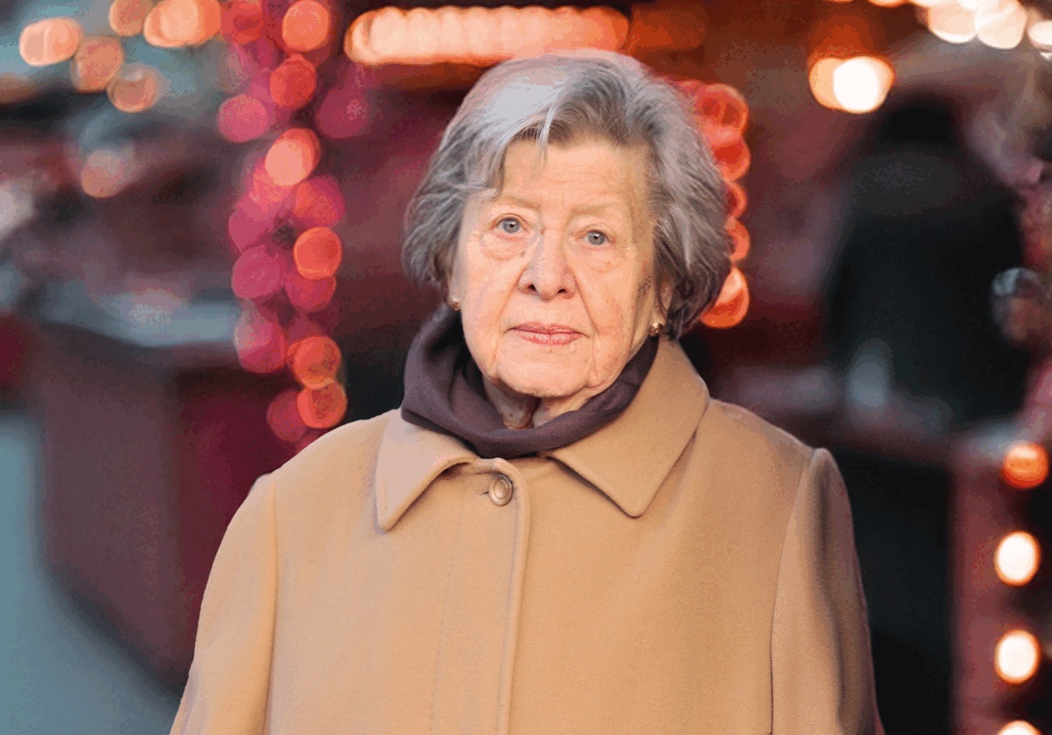 Eine ältere Dame mit grauen Haaren und Wintermantel. Sie schaut betrübt in Richtung Kamera. Im Hintergrund ist schemenhaft ein beleuchteter Weihnachtsmarkt zu erkennen.
