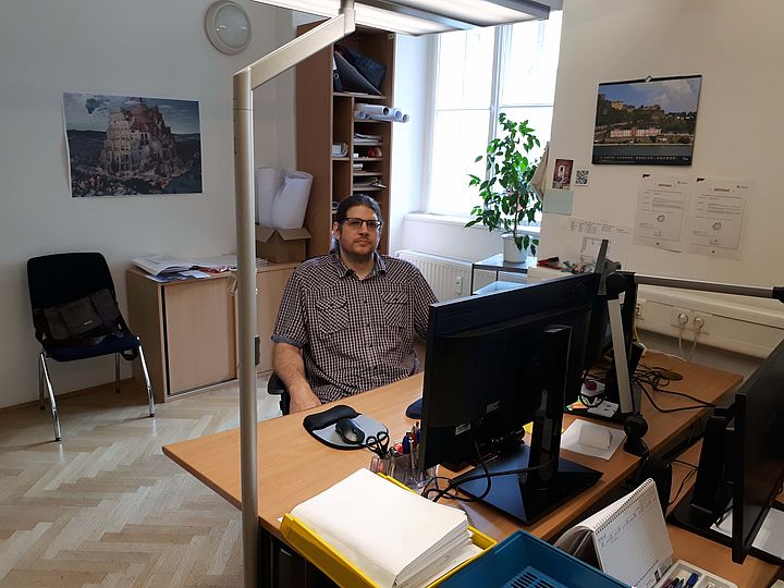 Ein Mann mit dunklen Haaren, Brille und leichtem Vollbart sitzt an einem Schreibtisch, neben dem Schreibtisch ist eine Stehleuchte platziert. Hinter ihm fällt durch ein großes Fenster Licht auf einen Kasten und einen Kunstdruck an der Wand.