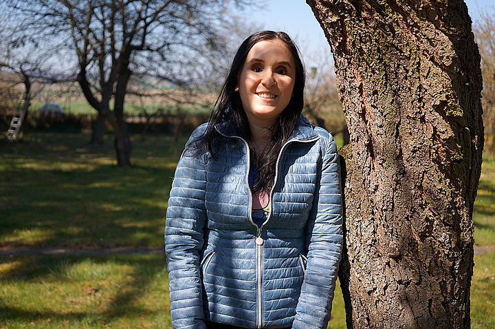 Eine junge Frau mit blauer Jacke und langen, glatten, dunklen Haaren lehnt in einem großen Garten an einem Baumstamm und lacht in die Kamera.