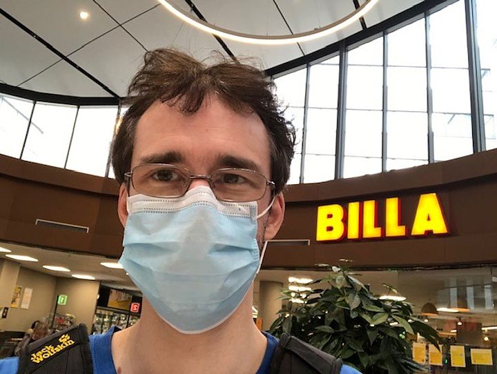 Ein junger Mann in Nahaufnahme mit Brille und Mund-Nasen-Schutz in einem Einkaufszentrum, hinter ihm ein großes BILLA-Schild
