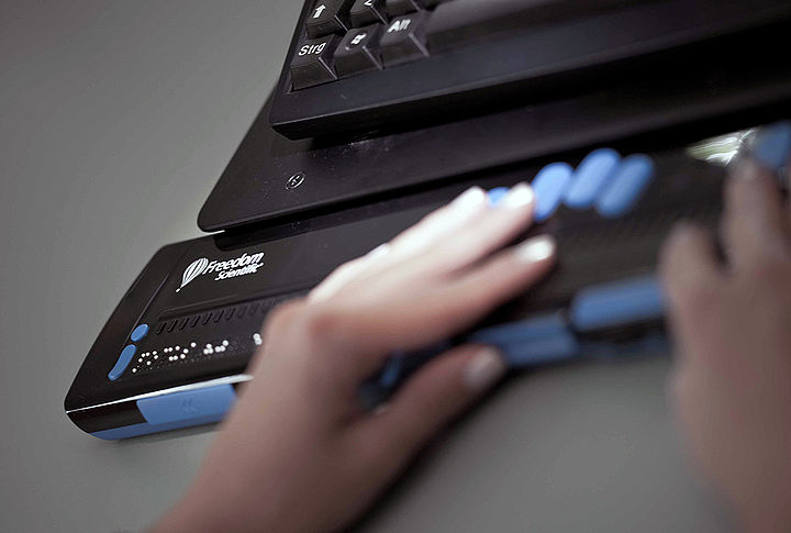 Nahaufnahme einer elektronischen Braillezeile vor einer Tastatur, über die ausgegebenen Braillepunkte gleiten die Finger zweier Hände