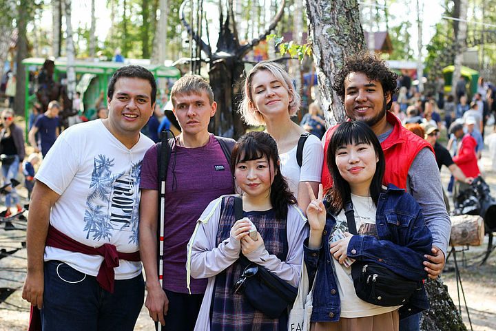 Drei junge Frauen und drei junge Männer stehen frohgemut für ein Gruppenfoto in einem belebten Park beisammen