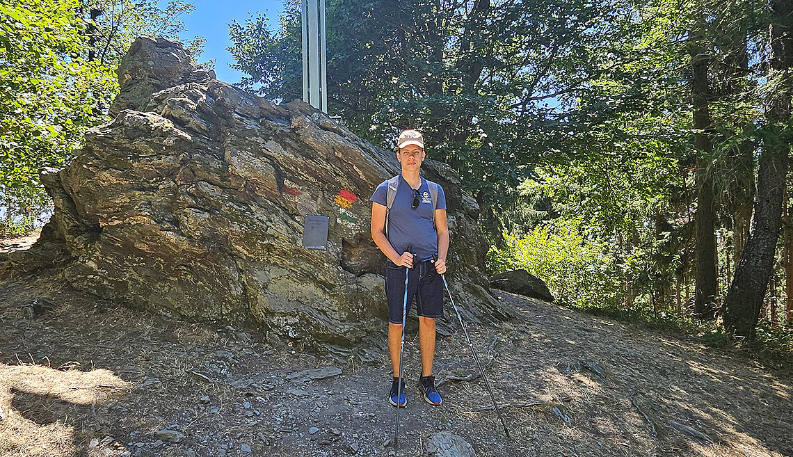 Mit Kappe und zwei Wanderstöcken steht ein junger Mann vor einem riesigen Felsbrocken am Höhepunkt einer Waldwanderstrecke.