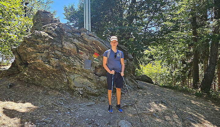 Mit Kappe und zwei Wanderstöcken steht ein junger Mann vor einem riesigen Felsbrocken am Höhepunkt einer Waldwanderstrecke.