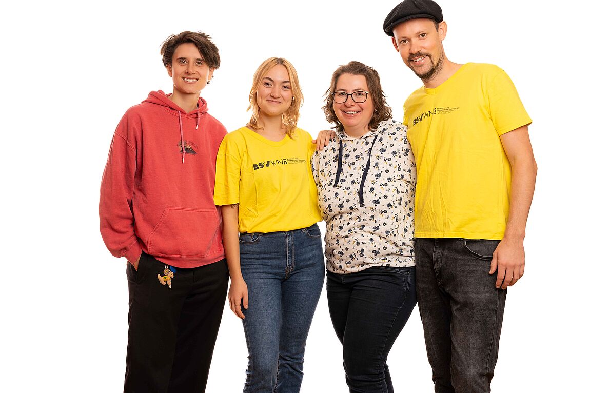 Zwei Frauen und zwei Männer, in dunklen Hosen und knalligfarbigen Shirts, stehen lachend für ein Teamfoto beisammen.