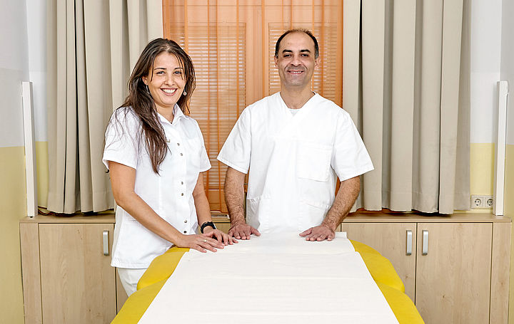 Eine Masseurin und ein Masseur stehen am Kopfende eines Massagetisches und lachen in die Kamera