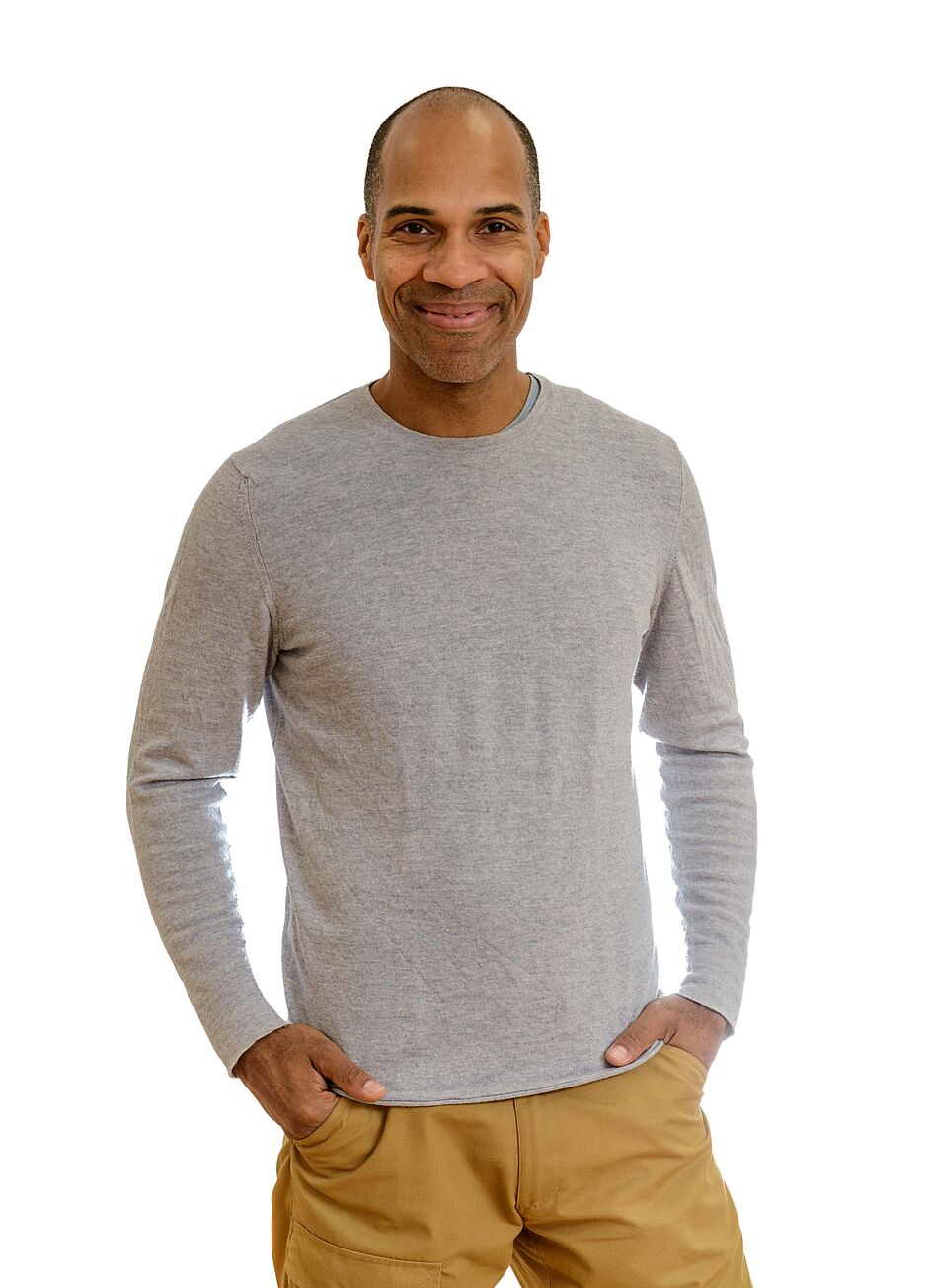 Ein Mann lächelt in die Kamera, trägt ein graues Shirt und beige Hosen, vor einem weißen Hintergrund.