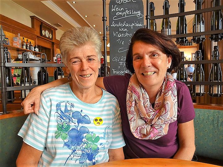 Zwei fröhlich lächelnde Frauen an einem Tisch in einer Gaststube, eine legt ihren Arm locker um die Schulter der anderen