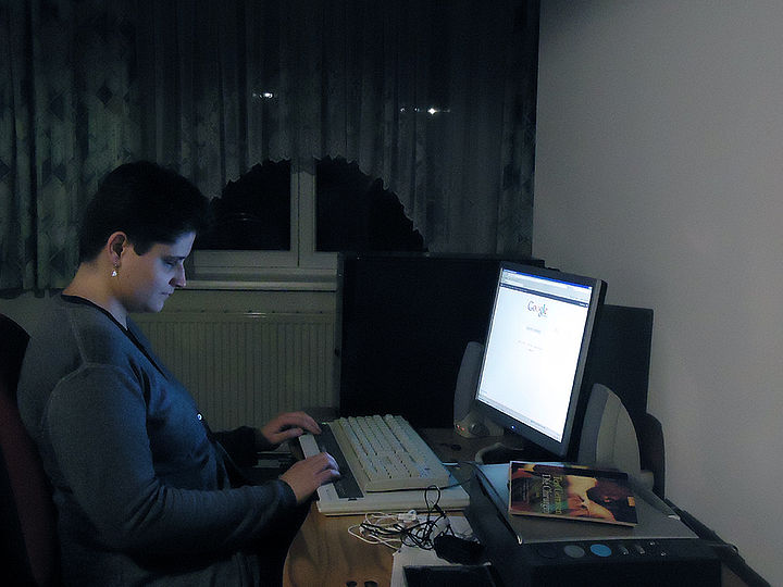 Eine junge Frau sitzt abends in einem dunklen Heimbüro vor der Computertastatur mit angeschlossener Braillezeile, der Monitor wirft Licht in den Raum