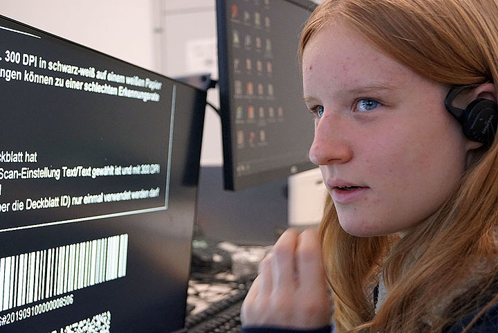 Eine junge Frau mit langen blonden Haaren vor einem Computerbildschirm, im Ohr einen In-Ear-Kopfhörer