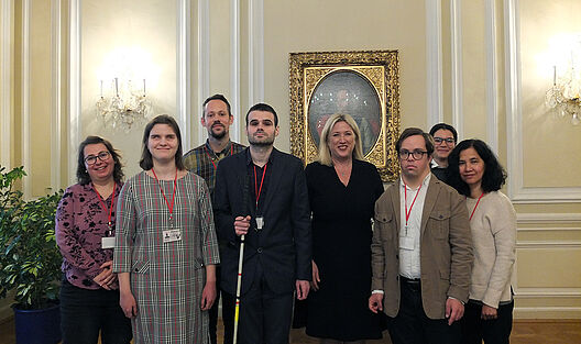 In einem prachtvollen Saal der britischen Botschaft steht eine Gruppe Teilnehmender mit zwei Betreuer*innen, während die Botschafterin zentral in der Mitte positioniert ist. Die aufwendig dekorierten Wände und Spiegel des Palais bilden den eindrucksvollen Hintergrund.