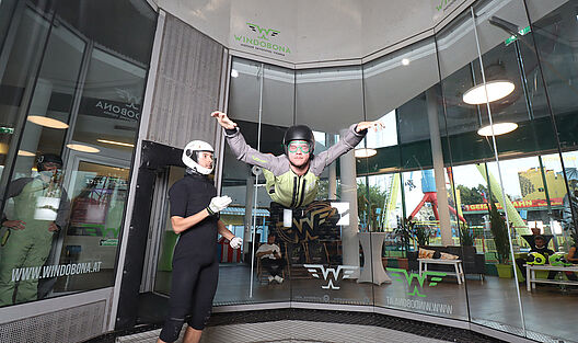 Ein junger Mann fliegt in einem Fluganzug gekleidet, mit ausgestreckten Händen und Füßen im Windkanal circa 2 Meter über dem Boden, dem sogenannten Skydiving im Prater
