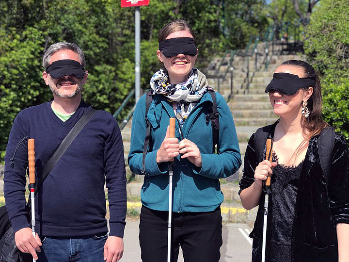 Ein Mann und zwei Frauen stehen vor Stufen und Büschen nebeneinander, alle haben die Augen mit Augenbinden verdeckt und halten je einen ausgeklappten Weißen Stock in Händen.