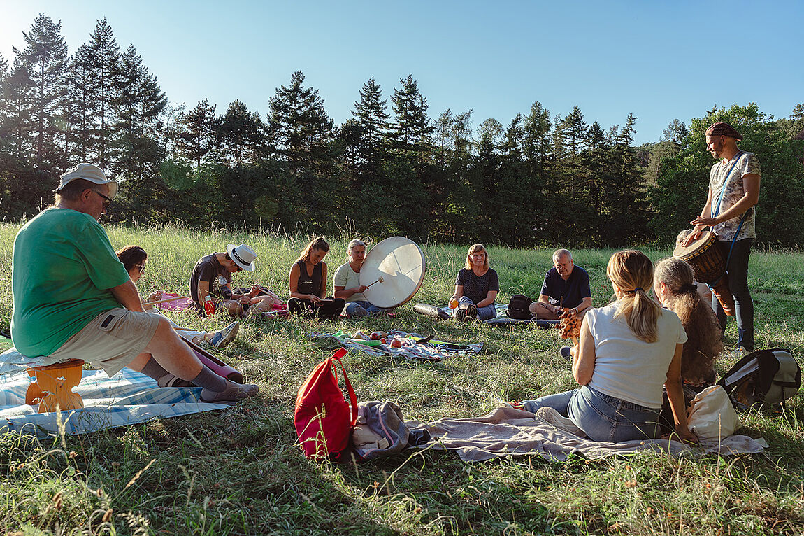 Ein knappes Dutzend Personen sitzen im Kreis auf einer Wiese, ein Mann steht mit einer Trommel am Bildrand und leitet die Gruppe an.