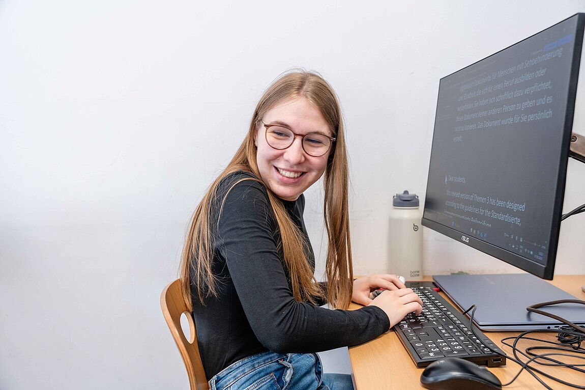 Eine lachende junge Frau mit Brille vor einer Tastatur und einem Bildschirm mit vergrößerten Inhalten.