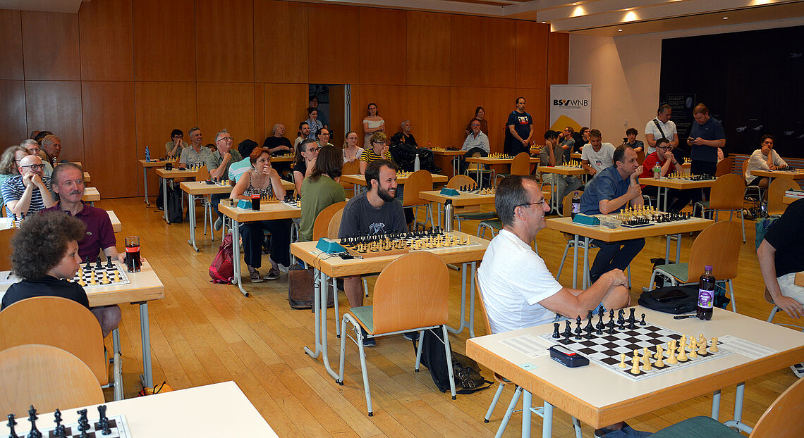 In einem großen Saal stehen in gleichen Abständen viele Tische, auf denen Schachutensilien vorbereitet sind. An mehreren Tischen sitzen schon Turnierteilnehmende und sind bereit für den Start.