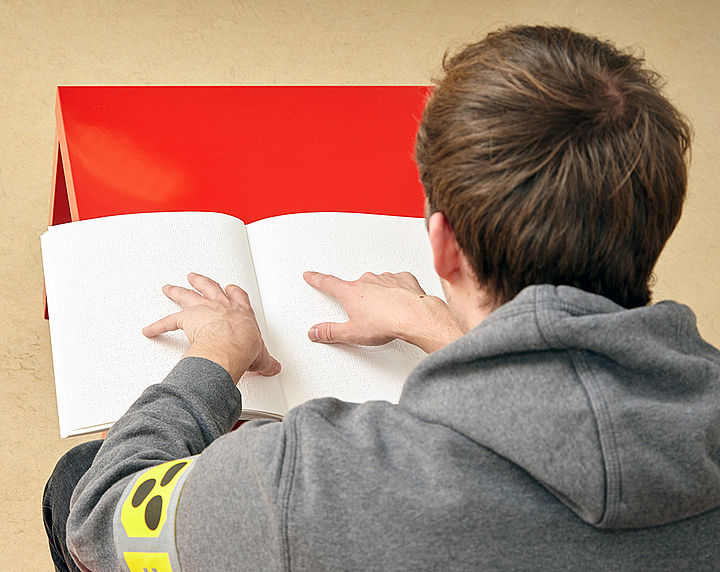 Auf einem roten Tisch liegt ein dickes weißes Buch, man sieht die Arme eines Mannes mit einer Blindenschleife links, der Zeigefinger der rechten Hand streicht über die erhabenen Braillepunkte auf der rechten Seite des Buches