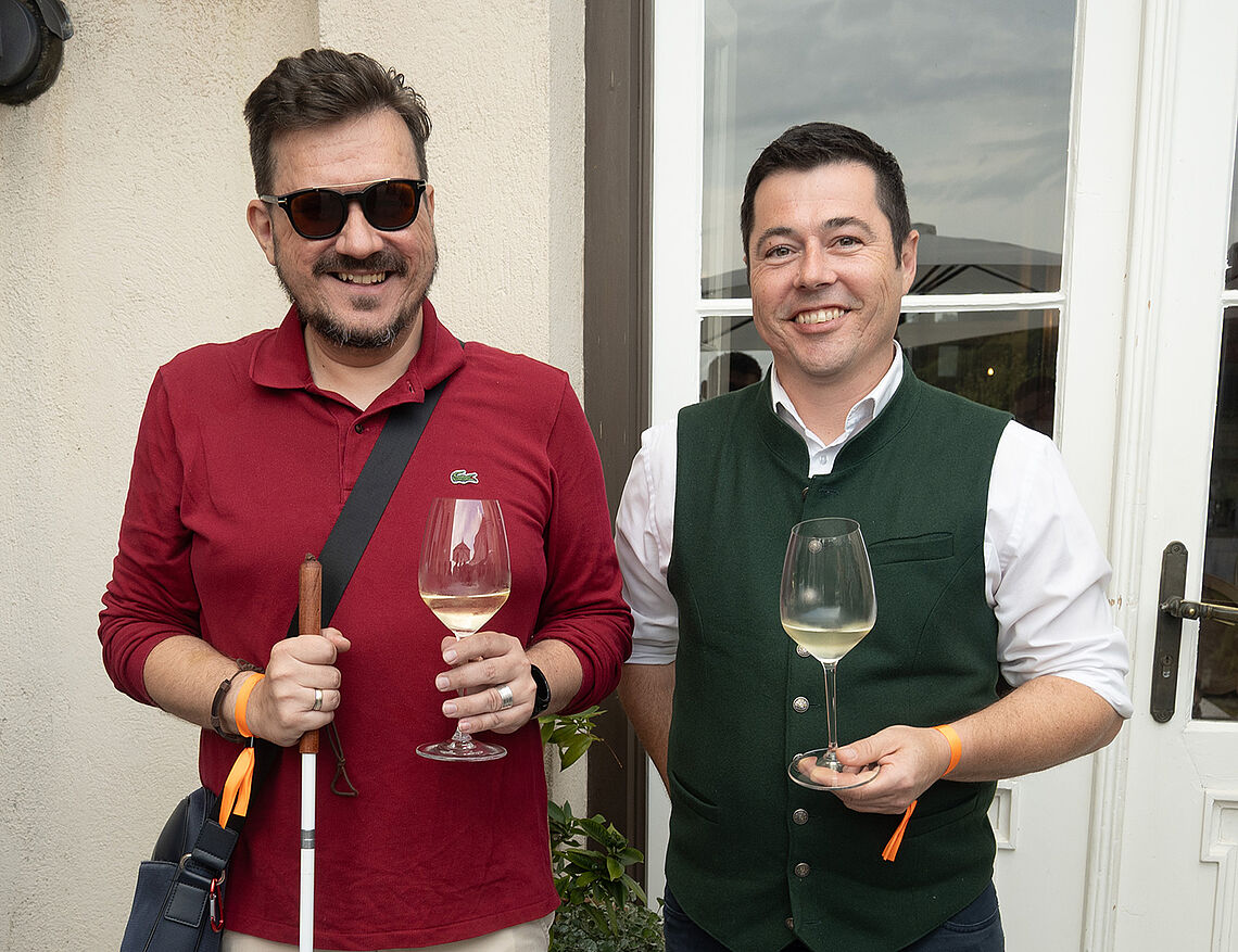 Zwei lachende Männer, beide mit einem Glas Weißwein, stehen vor einer rustikalen Außenmauer. Einer der beiden Männer hat zusätzlich einen Weißen Stock in der anderen Hand.