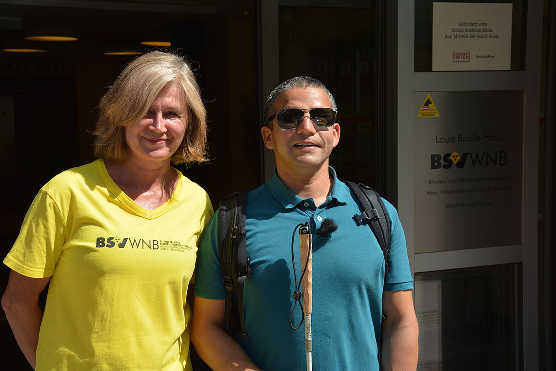 Eine Frau in gelbem T-Shirt und ein Mann mit Sonnenbrille und weißem Stock stehen im Sonnenschein vor dem Eingang des Louis Braille Hauses