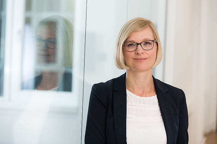 Portraitfoto von Julia Moser, sie trägt eine Brille, ihr Gesicht und ihre blonden Haare spiegeln sich in einer Glasscheibe neben ihr.
