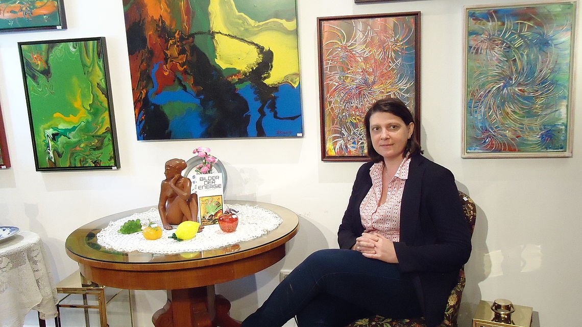 Barbara Probst sitzt an einem kleinen, runden Tisch mit Obstdeko, einer kleinen Frauenstatue und Blumen vor einer Wand mit farbenkräftigen Gemälden in verschiedenen Größen