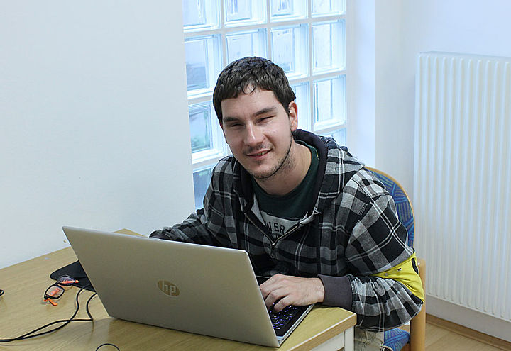 Ein junger Mann mit gelber Schleife am Arm tippt in einen Laptop, der auf einem kleinen Tisch vor ihm steht