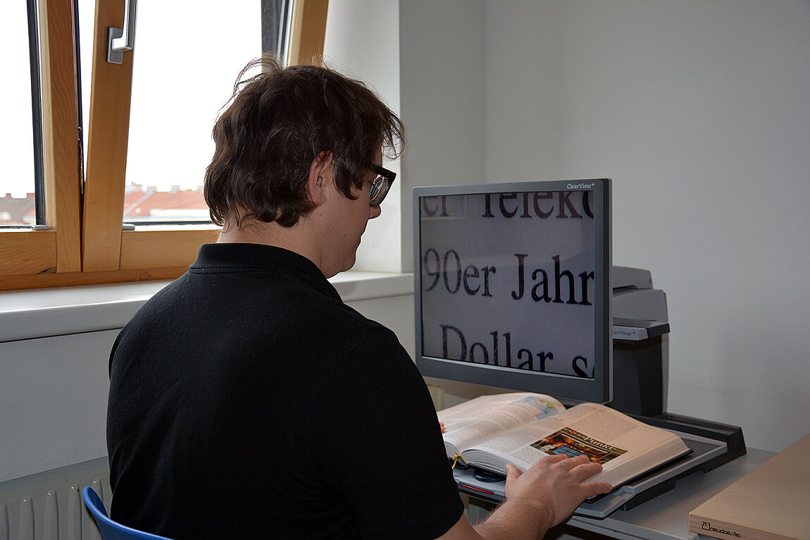 Ein junger Mann sitzt vor einem Monitor mit stark vergrößerter Anzeige eines Textes aus einem Buch, das unter dem Monitor (mit eingebauter Kamera) auf einer Ablagefläche aufgeschlagen liegt.