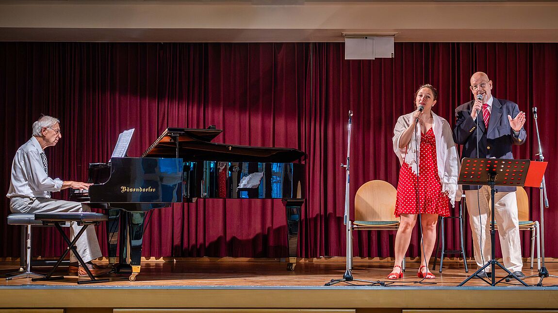 Mann spielt Klavier, Frau und Mann singen mit Mikrofonen vor rotem Bühnenvorhang