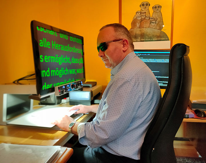 Ein Mann in blauem Hemd und mit Sonnenbrille sitzt auf einem Bürosessel vor einem großen Bildschirm, der darunter platzierten Text stark vergrößert darstellt.